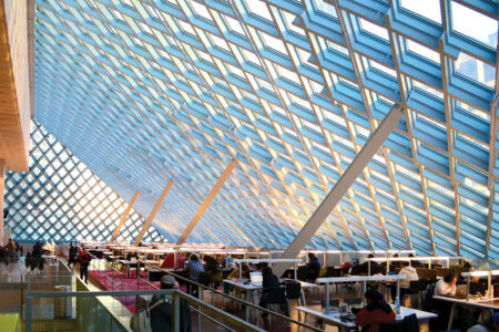 A modern library interior with a unique geometric glass ceiling allowing natural light. People are seated at tables reading and using laptops. The space has a bright and open atmosphere with a mixture of natural and artificial lighting.