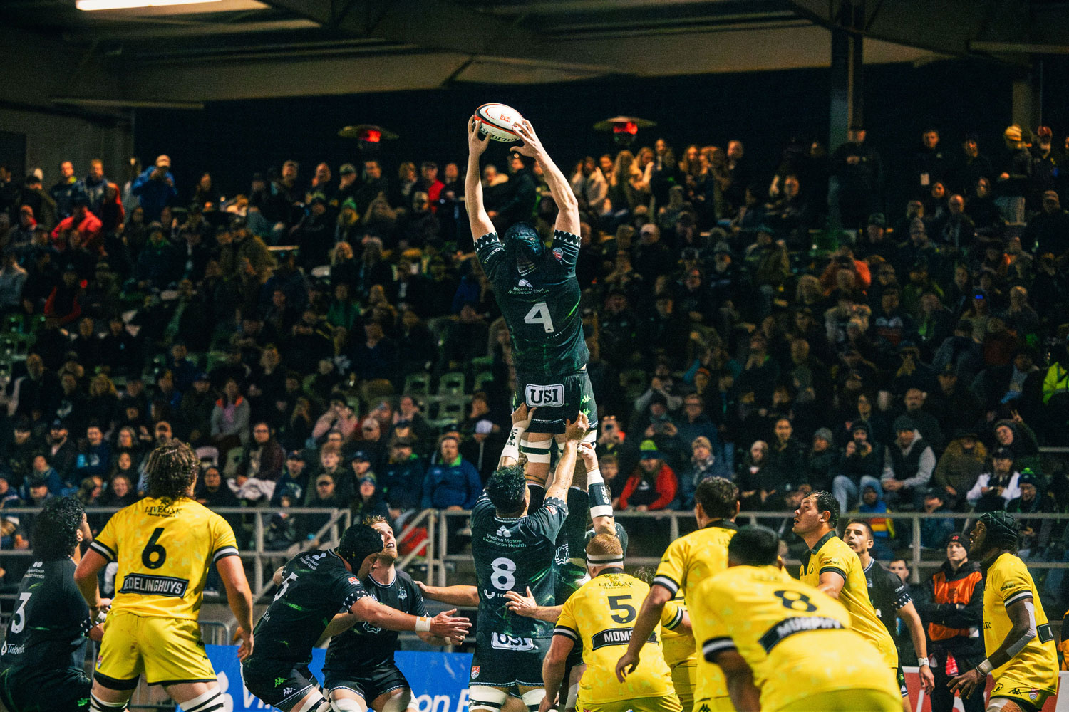 Amidst the cheers of a packed stadium, a player from the team in black leaps high to catch the ball during a lineout, surrounded by teammates and opponents in yellow. This thrilling rugby match captures the spirit of Seattle sports at its finest.