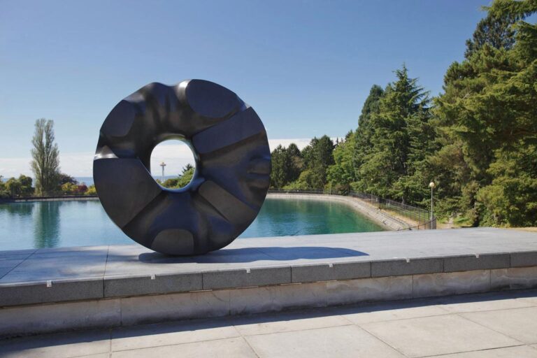 A large, circular black sculpture with abstract shapes stands on a platform overlooking a body of water. Trees and a pathway line the water's edge, with a clear blue sky in the background. A distant tower is visible beyond the trees.