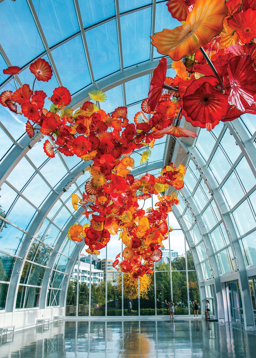 A large glass ceiling installation features vibrant orange, red, and yellow flower-shaped sculptures in a bright, spacious atrium with reflective flooring. The arched windows reveal a view of trees and buildings outside.