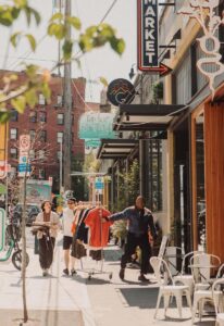Busy city sidewalk with people walking and browsing outdoor clothing racks near storefronts. Sunlight casts shadows, and street signs and bikes are visible among urban buildings and shop signs.