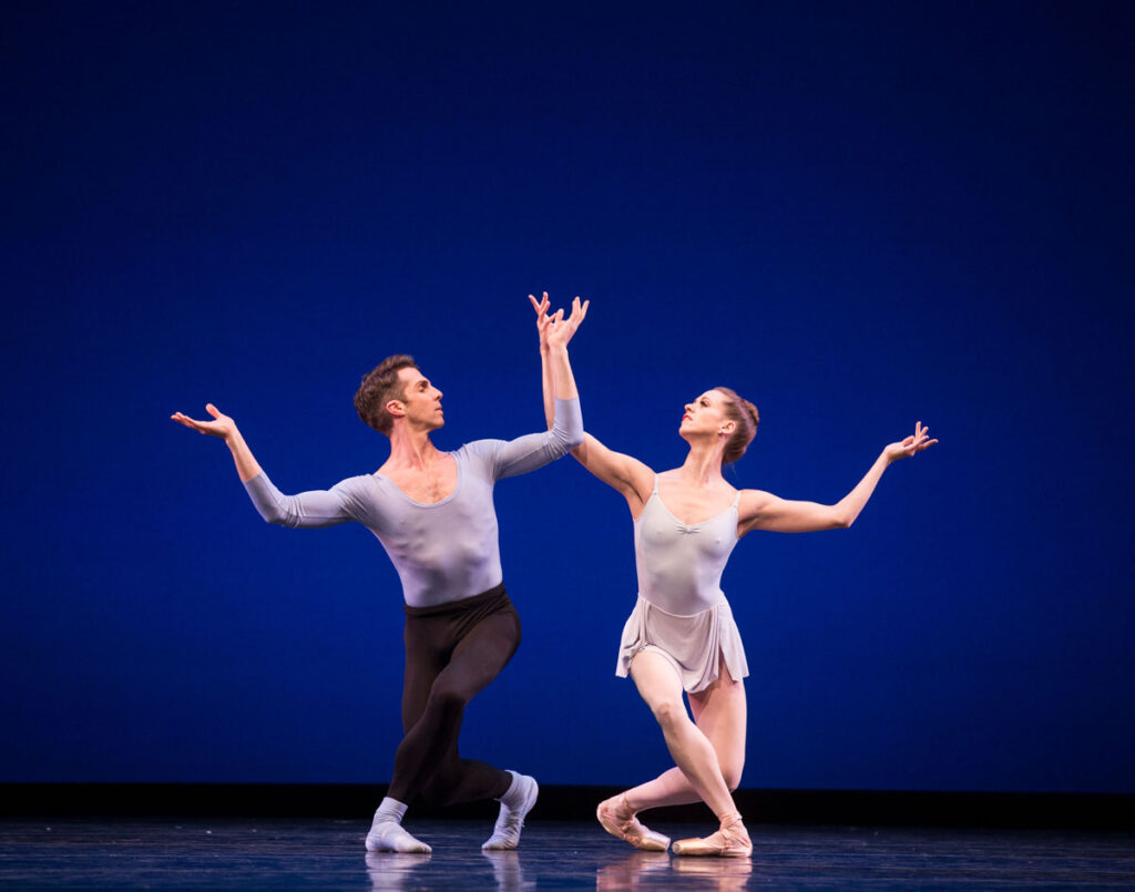 Two ballet dancers, a man and a woman, perform on stage against a deep blue background. Both are kneeling with one arm gracefully raised, wearing form-fitting dance attire.