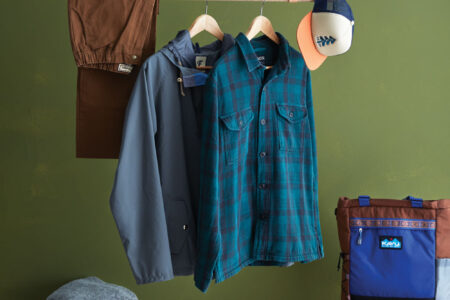 Two long-sleeve blue shirts hang next to folded brown pants and trucker hat on a clothes rack above a folded grey sweater, brown hiking boots, and a tote bag.