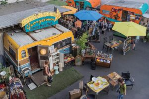 A colorful outdoor market with vintage trailers, clothing racks, furniture, and shoppers browsing under large umbrellas at the Georgetown Trailer Park Mall.