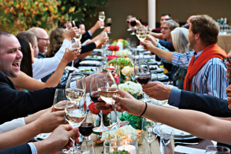a photo of a large group of people sitting at a long table raising their glasses to cheers.