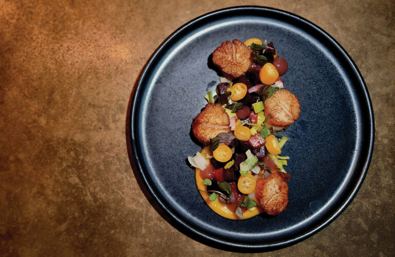 A plate of seared scallops arranged with sliced cherry tomatoes, diced vegetables, and greens on a dark plate, set against a textured brown background.