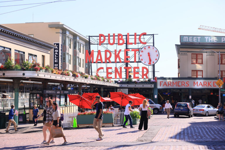 People walk through a bustling square in front of the iconic "Public Market Center" sign, one of the top things to do in Seattle. Red umbrellas are set up in a seating area, and various market stalls are visible. Buildings surround the scene under a clear blue sky.