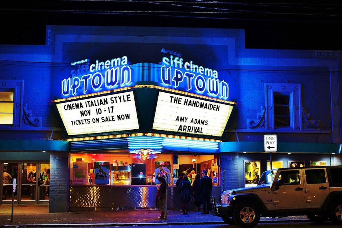 A vibrant night scene of the SIFF Cinema Uptown with bright neon lights. The marquee displays movie titles: "The Handmaiden," "Amy Adams: Arrival," and "Cinema Italian Style, Nov 10-17." People and a white SUV are visible in front.