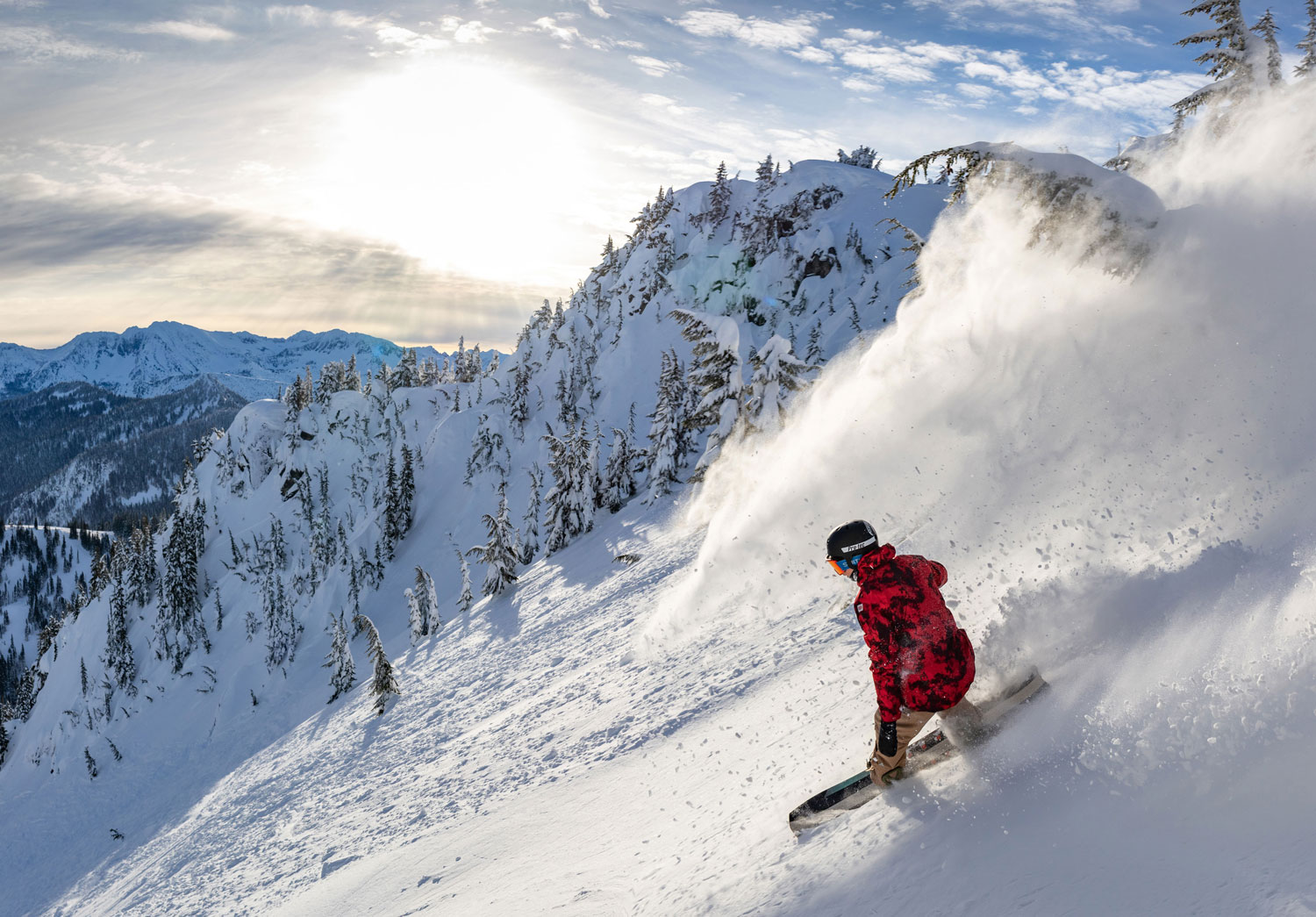 A snowboarder in a red jacket rides down a snow-covered mountain slope, surrounded by snow-capped trees. The sun casts a bright glow in the blue sky, highlighting the mountain ranges in the background. Snow swirls behind the snowboarder.