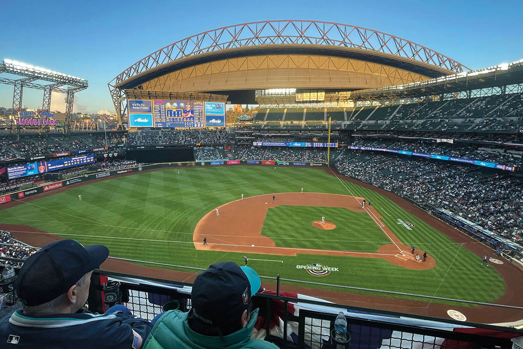 A wide-angle view of a Seattle sports stadium during a baseball game features a lush green field, players, and a partially filled audience. The stadium's retractable roof is open, with LED screens displaying game information under the clear blue sky.