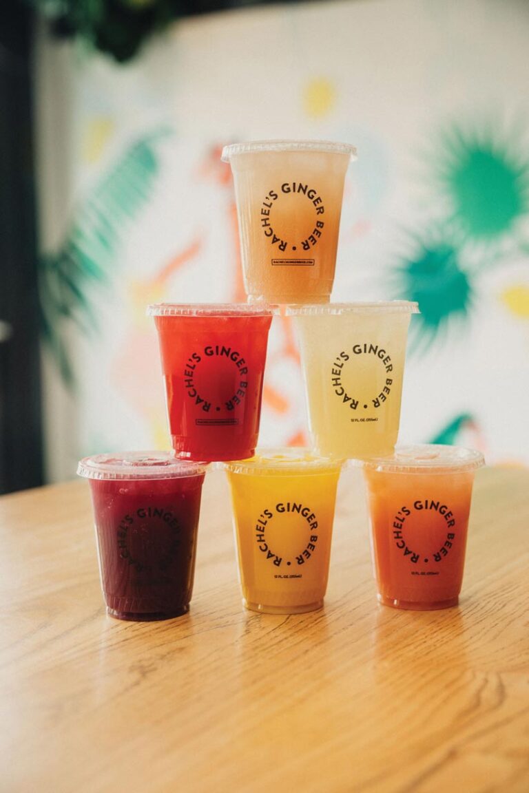 A pyramid of six colorful drink cups with transparent lids on a wooden table. Each cup has "Chef's Ginger Beer" printed on it. The drinks vary in color from red to yellow, against a vibrant, blurred background.