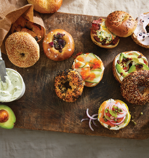 A variety of bagels on a wooden board, topped with cream cheese, avocado, smoked salmon, and vegetables. Nearby, there's a small cup of herbed cream cheese and a whole avocado. A paper bag holds more bagels.