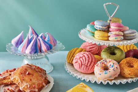 Assorted colorful pastries displayed on a teal tablecloth. Includes meringues, macarons, donuts, cookies, a cake slice, and fruit-filled pastries on decorative cake stands. Plates and a fork are set nearby.