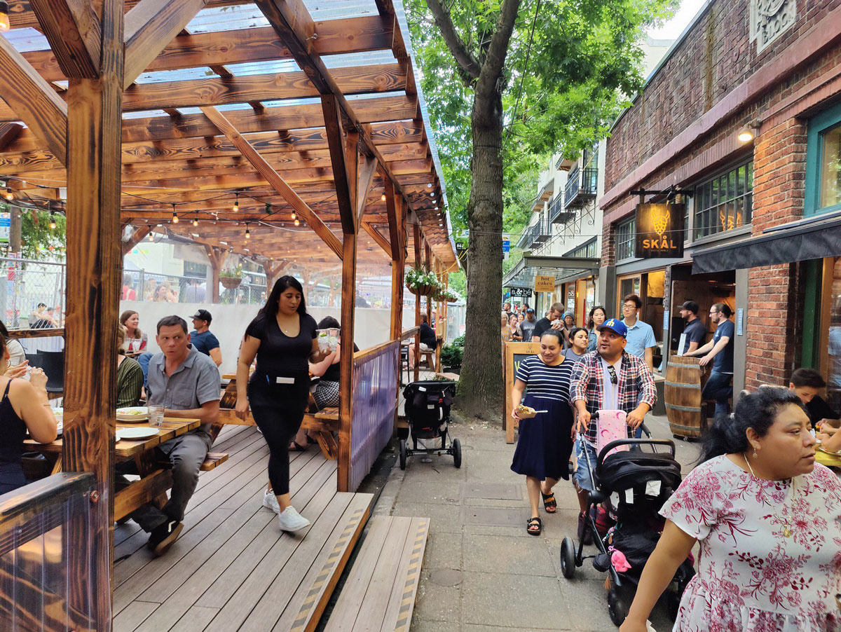 People dine at outdoor tables under a wooden pergola, while others walk past on a lively sidewalk lined with trees and storefronts. Some people push strollers, and a server walks toward the diners.