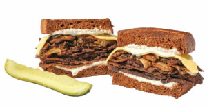 A sandwich with layers of thinly sliced meat, cheese, and sauce between dark rye bread slices, served with a dill pickle wedge on the side.