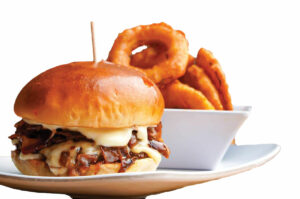 A burger with caramelized onions and melted cheese is served on a plate next to a bowl of crispy onion rings. The burger is topped with a glossy bun and secured with a toothpick.