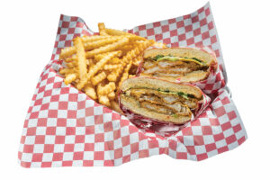 A chicken sandwich cut in half, filled with crispy chicken, lettuce, and sauce, is placed on red and white checkered paper next to a serving of crinkle-cut fries.