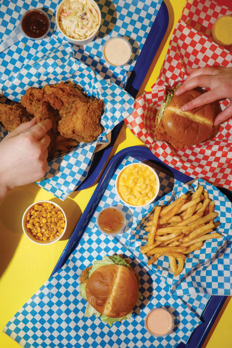 Trays of fried chicken, burgers, fries, macaroni and cheese, corn, coleslaw, and dipping sauces on blue and red checkered paper, with hands reaching for food on a yellow table.