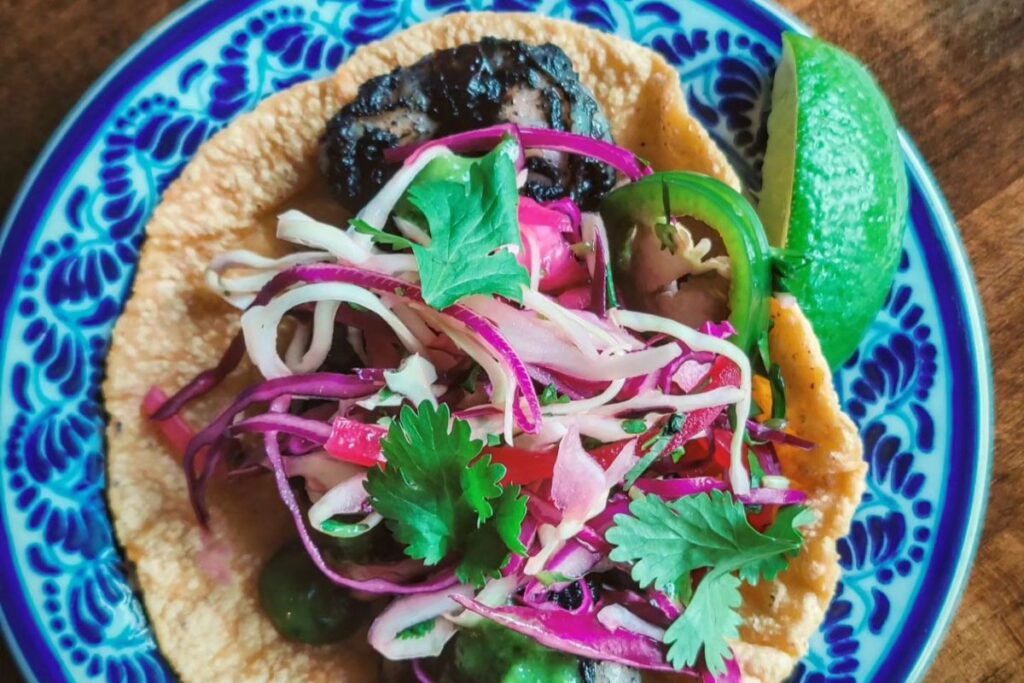 A corn tortilla topped with grilled meat, shredded red and green cabbage, cilantro, sliced jalapeño, and pickled vegetables, served on a blue patterned plate with a lime wedge.
