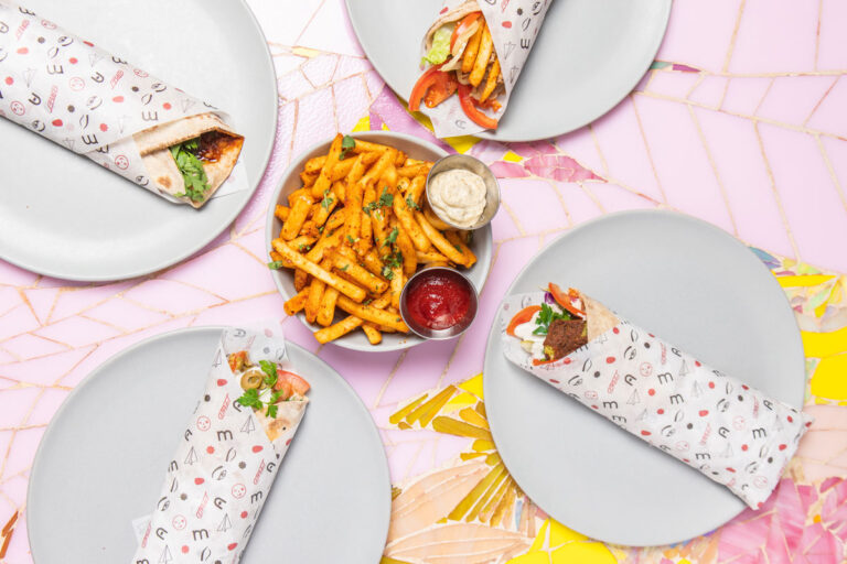 Four wrapped sandwiches on gray plates surround a serving of seasoned French fries with ketchup and a creamy dip, all placed on a pink and yellow mosaic-tiled table.