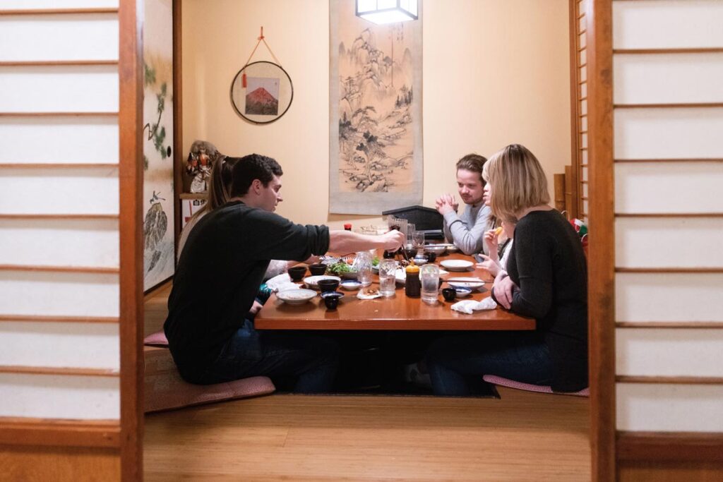 Three people sit around a low table in a traditional Japanese room, sharing a meal. The room features sliding doors, wall art, and a hanging decoration. The table is filled with various dishes and drinks.