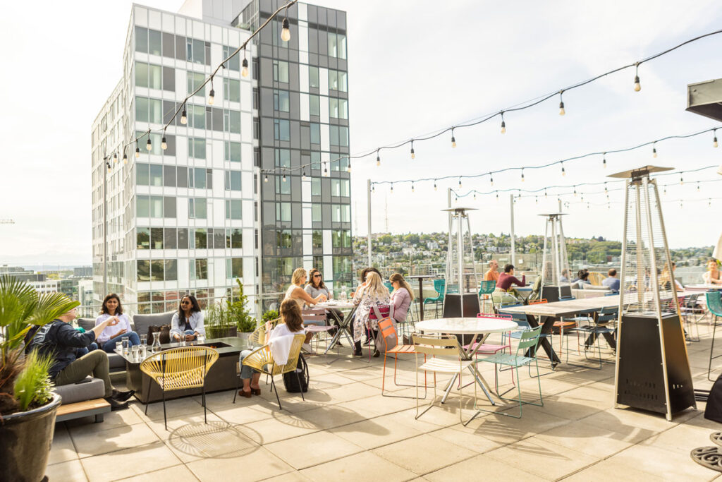 People sit and converse at tables on a sunny rooftop patio, surrounded by string lights, outdoor heaters, and city buildings in the background. Some enjoy drinks, while others relax and chat in small groups.