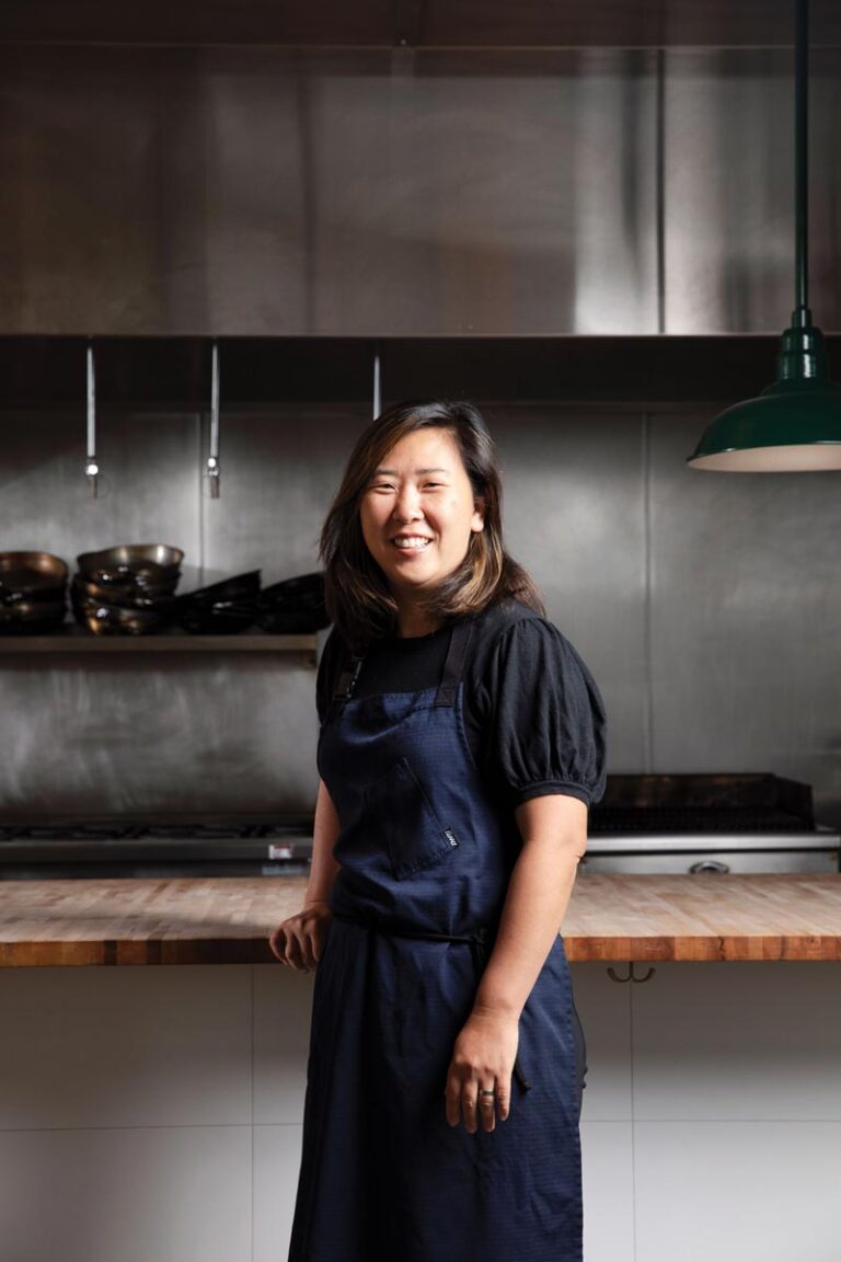 A woman wearing a dark apron and black shirt stands in a modern, stainless-steel kitchen, smiling at the camera. Shelves with bowls and a green hanging lamp are visible in the background.