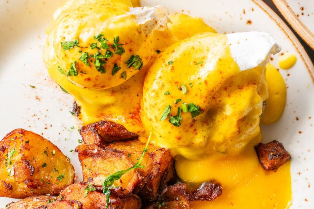 Close-up of eggs Benedict topped with hollandaise sauce and fresh herbs, served with golden crispy roasted potatoes on a white plate.
