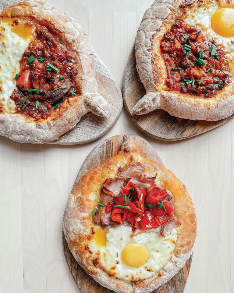 Three oval-shaped breads with thick crusts on wooden boards. Two are topped with tomato sauce and chives, while the third has a baked egg, cheese, bacon, and red peppers. All are displayed on a light wood surface.