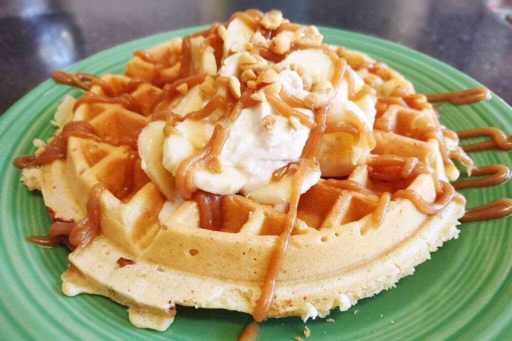 A plate with a thick waffle topped with whipped cream, banana slices, chopped nuts, and drizzled with caramel sauce, served on a green dish.