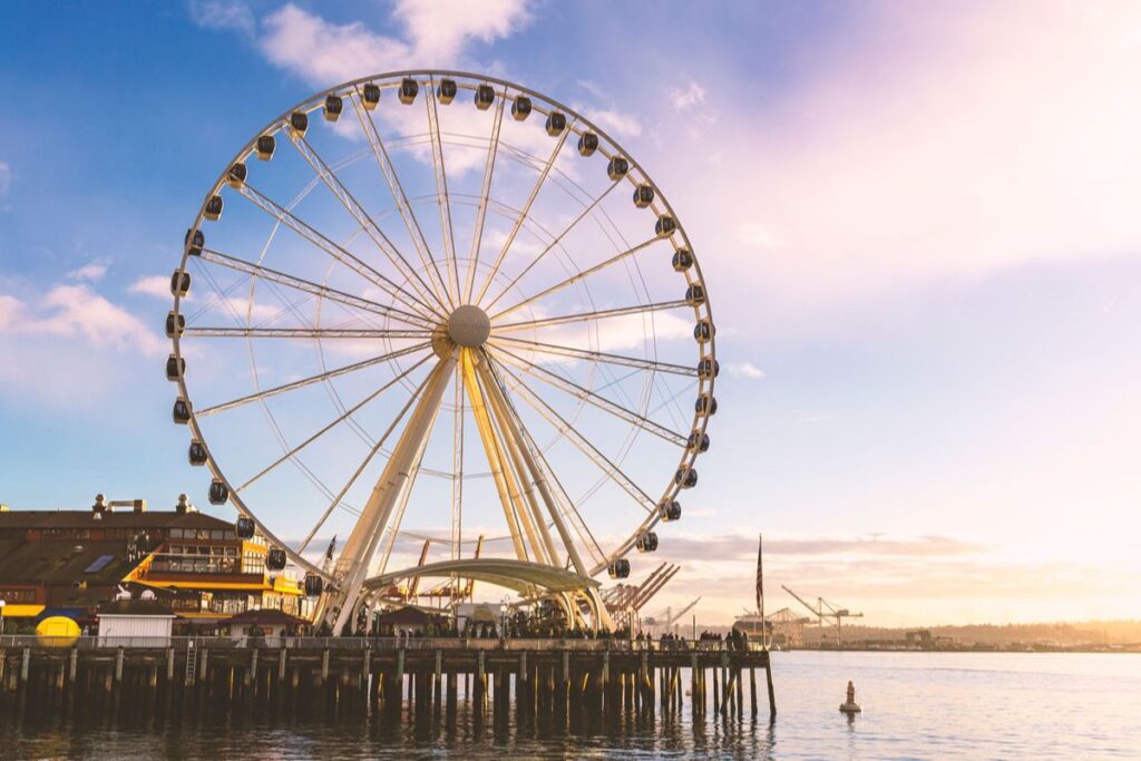 A large Ferris wheel stands on a pier over calm water at sunset, with soft clouds in the sky and buildings visible on the left side of the image.