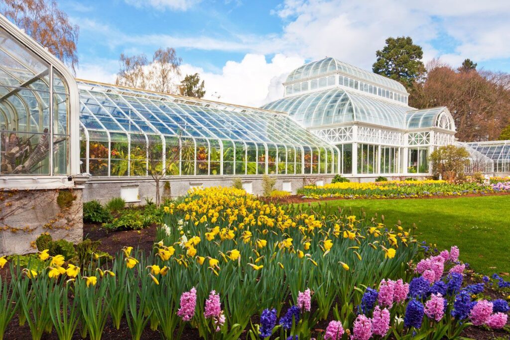 A large glass conservatory with arched roofs stands behind colorful flowerbeds filled with yellow daffodils and pink hyacinths, surrounded by green lawns and trees under a partly cloudy sky.