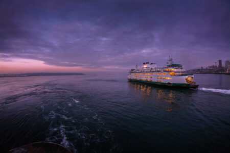 A photo of a Washington State Ferry moving across Elliott Bay. The sunset sky is purple and pink.