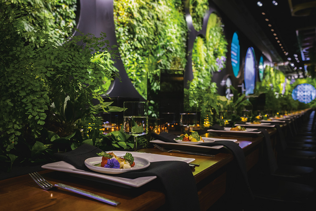 A photo of a dining experience within Climate pPledge Arena. Table settings with white plates and black napkins fill the table set again the background of the living wall within the arena.