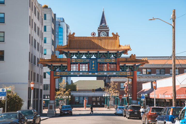 A vibrant Chinatown gateway with intricate designs and a pagoda-style roof arches over a street lined with buildings. A clock tower is visible in the background, and a person walks across the street below a clear blue sky.