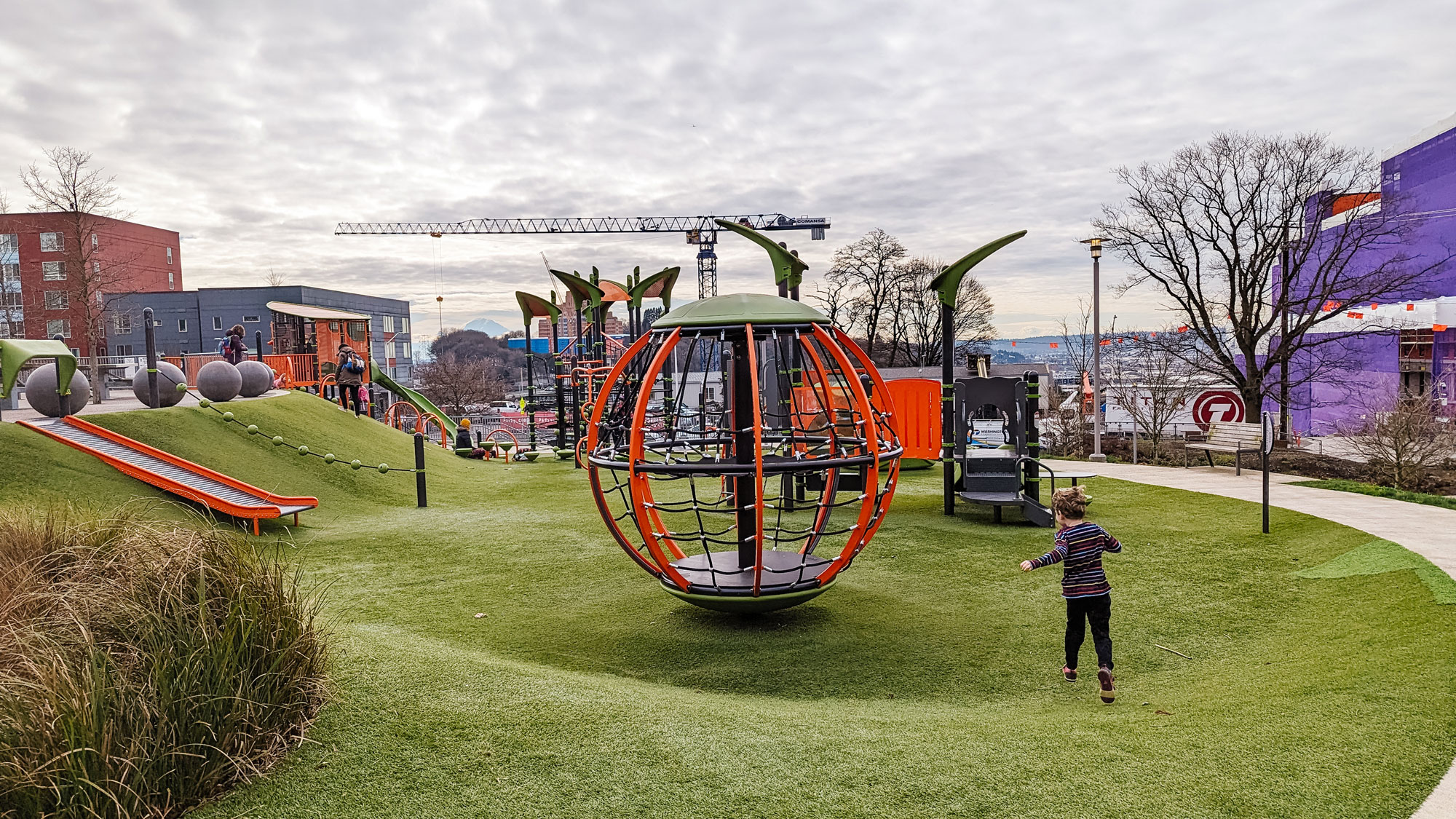 A large patch of green turf is the base of a colorful urban playground. An orb climbing structure is in the center of the image. It has orange metal bars, black netting, and a green top. A white child with short blonde hair wearing a blue, white, and red striped shirt and black pants runs towards the orb structure. On the left of the photo is an orange and silver metal slide coming out of the hillside. Other play structures with orange, green, and grey materials are in the background.
