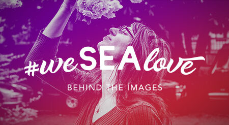 A woman stands outdoors, holding flowers above her head. The image has a pink and purple gradient overlay with the hashtag "#weSEAlove" and the text "BEHIND THE IMAGES" written on it.