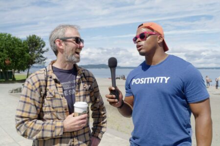 Two men are talking outside near a beach. One holds a microphone and wears a blue shirt with "POSITIVITY" written on it and red sunglasses. The other man wears glasses and a plaid shirt while holding a coffee cup. A few people are visible in the background.
