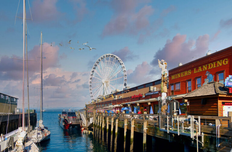 A waterfront scene with a Ferris wheel near a pier lined with shops and restaurants. A sailboat is docked nearby, with seagulls flying overhead. The sky is partly cloudy, and the water reflects the vibrant colors of the buildings and sky.