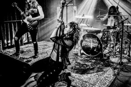 A black-and-white image of a rock band performing on stage. The guitarist is playing passionately, the singer is kneeling while holding the microphone, and the drummer is energetically playing drums. Stage lights create dramatic beams.