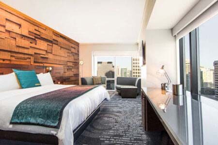 A modern hotel room features a large bed with a teal and white color scheme. The room has a textured wooden accent wall, a comfortable seating area by a window with city views, and a sleek desk with a lamp.