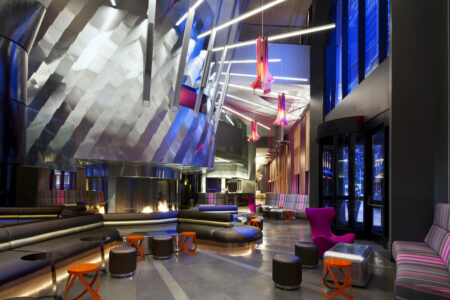 A modern hotel lobby with futuristic design features metallic walls, colorful hanging lights, and sleek seating including purple chairs, striped couches, and orange stools. Large windows let in natural light.