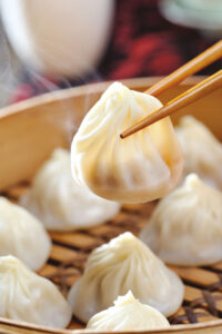 Chopsticks holding a steaming dumpling above a bamboo steamer filled with similar dumplings. The focus is on the lifted dumpling, showcasing its delicate folds and translucency.