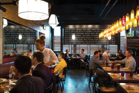 A busy restaurant interior with patrons dining at various tables. A server is attending to customers. The space is warmly lit with hanging lamps and features a wall displaying wine options. Guests are engaged in conversation, creating a lively atmosphere.