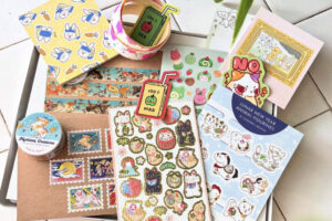 Assorted colorful stationery items, including animal stickers, washi tape, illustrated cards, stamps, and a small tin, neatly arranged on a white tiled surface. Designs feature cute animals and playful motifs.