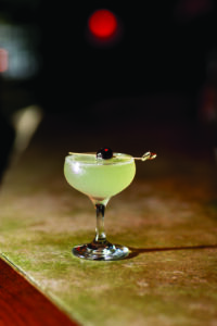 A cocktail in a coupe glass sits on a bar counter. The drink is garnished with a cherry skewered on a cocktail pick. The background is dimly lit, with a soft red light in the distance.