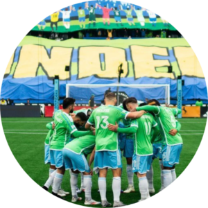 A soccer team in green uniforms huddles together on the field, with a large, colorful crowd display and banner in the stadium stands behind them.