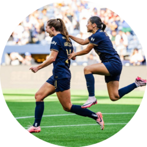 Two female soccer players in navy uniforms celebrate on the field; one is running while the other jumps joyfully behind her, reaching out with one hand. The background shows a crowd in the stands.
