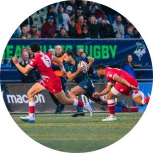 A rugby player in a dark uniform runs with the ball, pursued by two players in red uniforms on a field, while a crowd watches from the stands in the background.