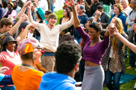 A diverse group of people dancing joyfully outdoors at a lively event. They are holding hands and smiling, surrounded by a cheering crowd. Bright clothing and a sense of celebration can be seen in the vibrant atmosphere.